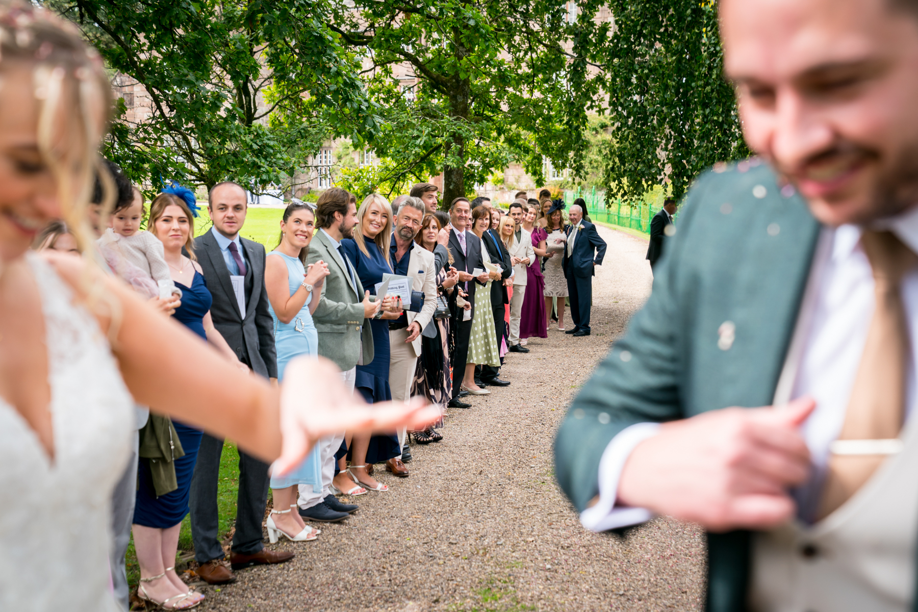 Wedding guests line a tree-shaded path beside newlyweds